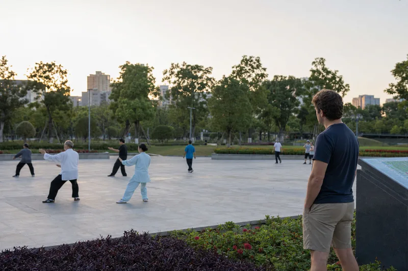 Traveler observing a morning exercise group from the edge of a Chinese public park